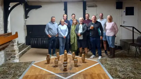 Perry St Social Club A group of people gathered at the end of a skittle alley with wooden skittles set up in front of them. Some people are holding drinks and they are all smiling at the camera. There is a highly patterned carpet either side of the skittle alley