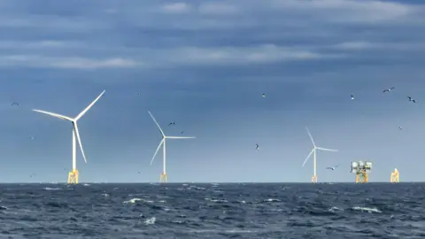 Neart na Gaoithe wind farm: There are three wind turbines on top of yellow platforms in the sea. A few birds fly over head.