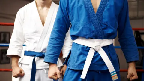 Getty Images Two people wearing Judo outfits. One is wearing a white judogi with a blue belt and the other is a blue judogi with a white belt.