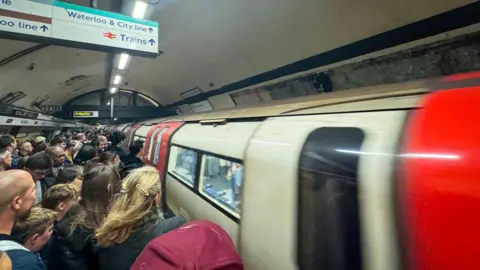 A tube platform packed with people waiting to get on to a waiting train. The doors of the train have not opened yet. Most of the people are facing away from the camera towards the train doors. 