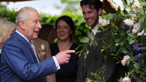 PA Media King Charles pointing towards some pink and purple flowers on the right. He is wearing a blue suit and is mid-speech. There are people standing behind him, on the right is a man with brown hair smiling at him.