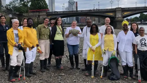 Thames21 A group of people stand on the foreshore of the River Thames in front of Vauxhall Bridge/ They are wearing wellies and some have litter pickers in their hands.