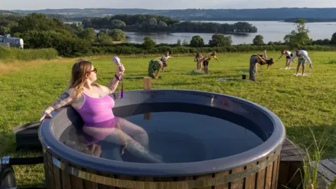 PA Media A woman in a pink swimsuit cools herself with a small handheld electric fan as she sits in a large circular plunge pool. Behind her in a field other people are stretching in various yoga poses while further in the distance the water and wooded islands of Chew Valley Lake is visible