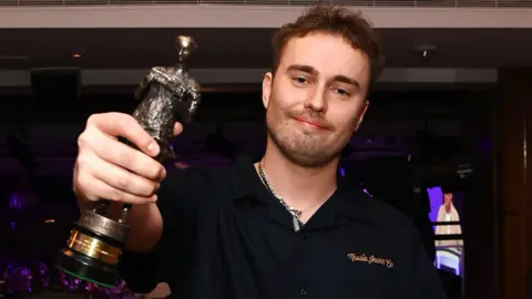 Getty Images Sam Fender wears a black shirt and holds a statuette to the camera as he smiles. In the background is the ceremony hall