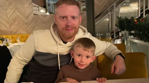 NCDS Michael Woods, 36, who has ginger hair and a short ginger beard, sits at a table in a restaurant with his arm around his son, six, who has short blonde hair. 