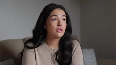 BBC / Jadzia Samuel A woman with dark hair and beige cardigan. She is sitting on a couch.