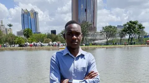 Kelvin Karume A photo of a man wearing a light blue shirt and navy pants standing in front of a lake with a city landscape in the background.