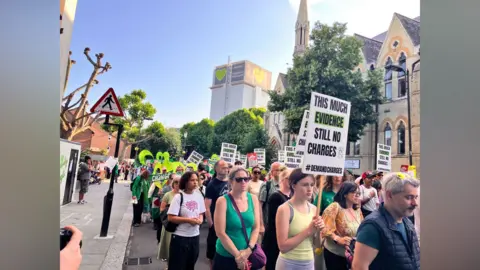A large group of people some holding placards walk down a street in a silent protest walk. The partly deconstructed Grenfell Tower is visible in the background.