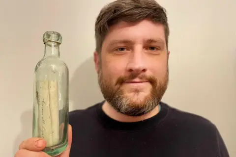 Ross Russell Ross is holding the bottle up near his face and smiling at the camera. He has a brown beard and brown hair and is wearing a dark blue jumper.