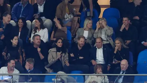 Getty Images Megan and Harry are seen sitting on blue stadium seats. She is wearing a tan jacket while he is wearing a dark button-up shirt and sitting with his arms crossed. There are people sitting on either side of them, behind and in front of them at the Allianz Stadium in Sydney.
