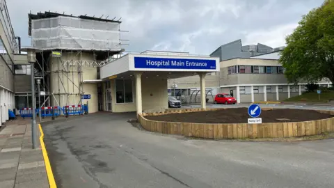 BBC Entrance to a hospital. A small roundabout is in front of the entrance. The building is white with a large blue entry sign over the door.
