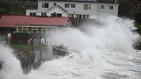 This picture shows powerful waves crashing against a coastal sea wall during rough weather. The water is surging high, creating large splashes and spray that dominate the foreground. Behind the sea wall, there are buildings very close to the shoreline—one with a red roof and another larger house. 