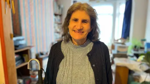 A woman wearing a navy jumper, she carries a cane and has grey hair