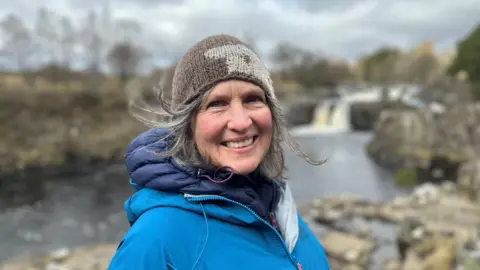 Sara Cox smiles at the camera. She is wearing a thick blue coat and brown woolly hat with a sheep's face designed on it. She has blue eyes and long greying hair flicking out from beneath her hat. Behind her are trees, a river and a small waterfall.