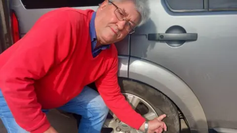 Conservative councillor Terry Mendies, wearing a red jumper, blue shirt and blue jeans, shows the damage to one of the tyres on his Land Rover Discovery