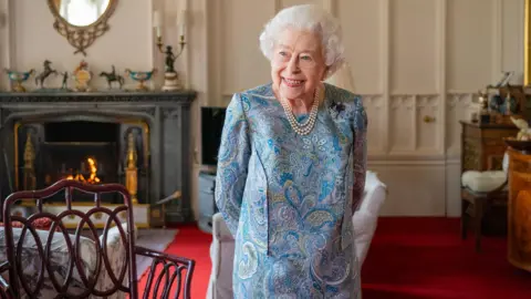 Queen Elizabeth II is standing inside a room Windsor Castle on April 28, 2022 wearing a blue dress and pearls. She is smiling