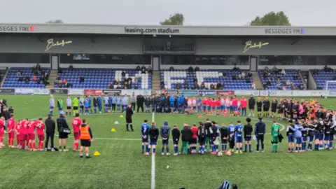 Football 4 Macclesfield Young footballers and adults connected to the team and summer camp stand in a circle on a pitch paying tribute to Brodan. They are wearing many different coloured kits and a referee appears to be standing in the middle of the circle.
