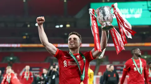 Reuters Jota is in a red Liverpool FC jersey holds up a trophy adorned with red and white ribbons. The player wears a green and white medal around their neck. In the background, other team members, spectators, and stadium lights are visible