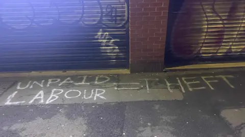 Graffiti sprayed in white paint on the ground outside one of the restaurants after it closed. The graffiti reads UNPAID LABOUR = THEFT