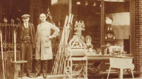 Historic England A sepia-coloured photo of two men, one in a flat cap and suit and other in an overall - standing by the cast iron gates with a variety of goods, including a mangle, outside and in the window.