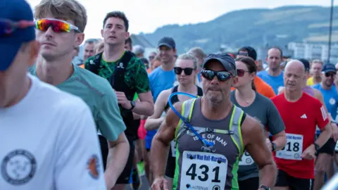 DAVE BELL /DKPHOTOGRAPHYIOM A group of people running with numbers on their torsos. One man wearing a cap and blue sunglasses runs past, you can see the hill in the background in Ramsey.