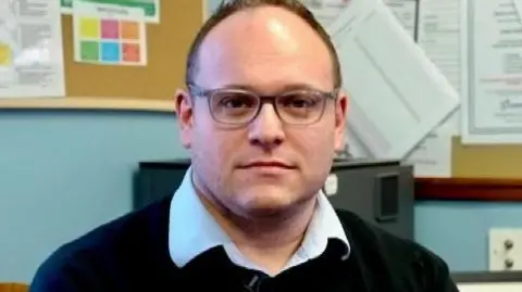 Dr Jamie Green in his surgery. He is wearing a light-coloured open-necked shirt over a dark sweater. He has short hair and glasses. Behind him is a pinboard and the back of a computer monitor.