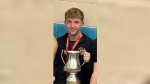 Supplied Cain Pears is smiling at the camera as he holds a silver trophy cup. He has short blonde hair and is wearing a black sweatshirt.
