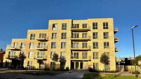 Danks Badnell Architects Ltd A modern yellow brick five-storey building with balconies to the front and sides and a concrete pathway leads to a main entrance at the front. Three small trees stand in front of the building