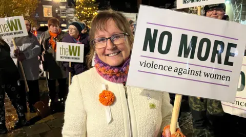 Woman with brown hair, white jumper, scarf, she is wearing glasses and holding a placard which reads, no more violence against women. There is a crowd behind her.