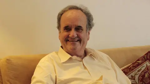 Sir Mark Tully sits on a sofa wearing a yellow shirt as lamp light falls on him 