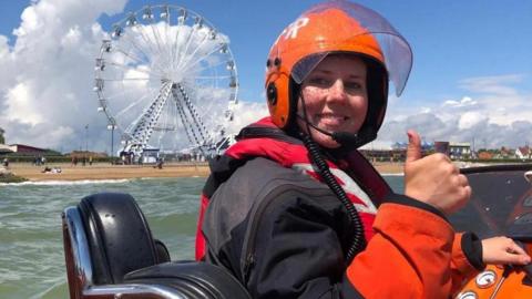 A smiling crew member Izzy on board a lifeboat giving a thumbs up to the camera, she is wearing a lifejacket and orange helmet. On the beach behind her is a Ferris wheel. 