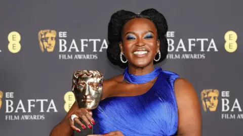 Wunmi Mosaku grins as she holds her Bafta award in front of the films awards backdrop. She wears a blue off shoulder dress and scarf.