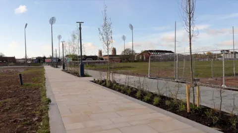 A light-coloured pathway runing through the middle of the shot, surrounded by green fields and fencing. In the distance are the floodlights of a cricket stadium