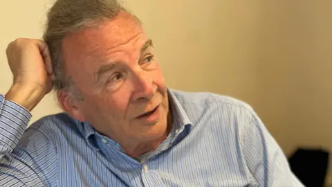 A man wearing a white and blue striped shirt can be seen leaning on his hand. He is looking off to his left as the photograph is taken. He has grey hair.