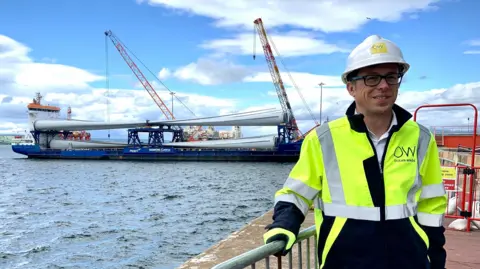 A man wearing a reflective vest and a helmet stands on the shore, and behind him in the distance is a cargo boat carrying a shipment of wind turbine blades