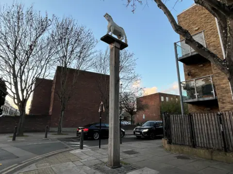 A large wooden carving of a white horse standing on a tall wooden plinth with buildings around it and two cars passing by on a road behind it