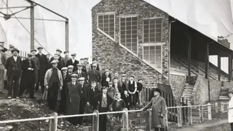 Workington AFC A black and white image of fans standing next to the stand at Borough Park in 1937, The stand is a brick construction with steps at the front leading up to the seats. The fans are standing on a mud bank. Their are a few boys near the front while the rest are men all wearing hats. 