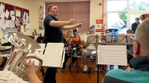 A brass band practising, with a conductor holding a white baton and a tuba player in the foreground.