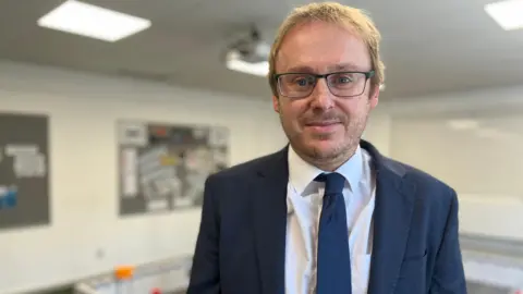Head of sixth form, Rob Shaw is smiling at the camera. He has fair hair and dark framed glasses. He is wearing a navy suit with a white shirt and navy blue tie. 