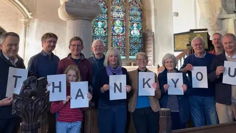 Copyright Hilary Rule/Ickleton Church 11 people standing in a church, in front of a window and above them are medieval frescoes. They are a mix of men and women plus one girl. The pulpit is to the right. Above them is an elaborate ironwork electric lamp. 