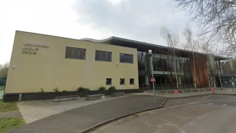 Google A large pale yellow building with windows adjoins a glass fronted building 