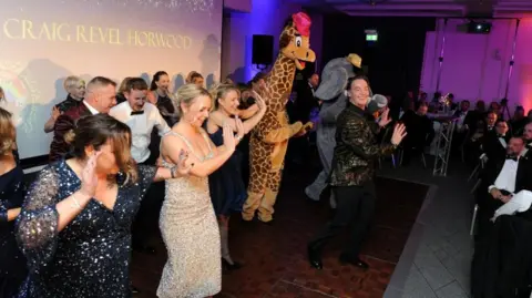 North West Anglia Hospitals’ Charity Craig Revel Horwood, wearing a gold spangled jacket over a black shirt at the front of a stage with a line of of men and women, plus behind him a mascot dressed as a giraffe and another dressed as an elephant. They are all dancing and laughing and following Revel Horwood's directions. In front are some tables with men in black tie. 