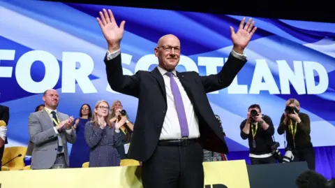 Getty Images John Swinney at an SNP conference, arms aloft, taking applause in front of a big sign reading "for Scotland"