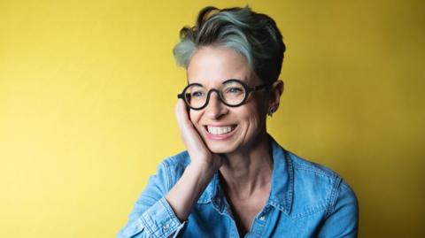Liz is wearing a denim shirt against a yellow wall. She has short, grey hair and is wearing black-rimmed glasses and is resting her chin on her hand.