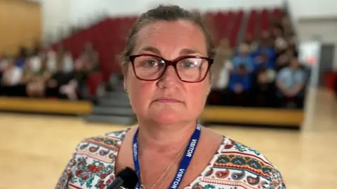 In dark-framed glasses and with her hair tied back, Keena Entwistle stands in a school hall looking at the camera with an audience of sixth form students seated behind her.