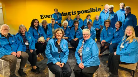 Twenty-two people and one dog sitting down, facing the camera and smiling, all wearing matching blue jackets 