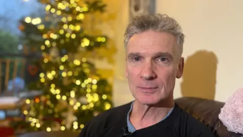 BBC A close-up of a man with silver hair sitting on a sofa and looking into the camera. The background is blurred but he is sat in a living room with a lit-up Christmas tree behind him.