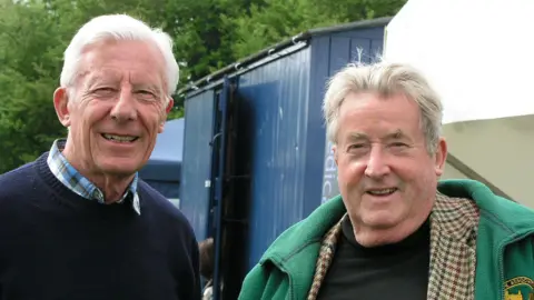 GWR Cyril Bleasdale stands on the left in a blue jumper and grey hair with colleague Sir William McAlpine in a green jacket in 2013. Both men are smiling and looking at the camera.