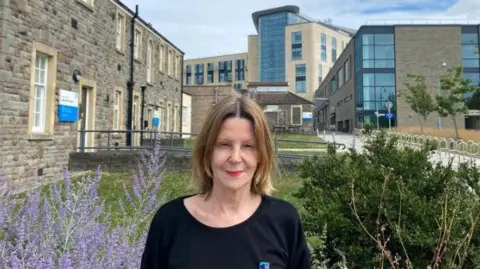Maria Kane is standing in a garden with Southmead Hospital's main building behind her. She has dark blonde shoulder-length hair and is wearing a black top.