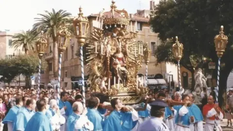 Phil Roberts A grainy colour photo of men from the Confraternity of Nettuno holding a statue of the Madonna on their shoulders on a street in Nettuno. The men are wearing white robes with light blue capes. Behind them are crowds of people, palm trees and buildings. 